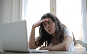 Eine junge Frau sitzt vor einem Laptop und schaut verzweifelt in die Ferne.