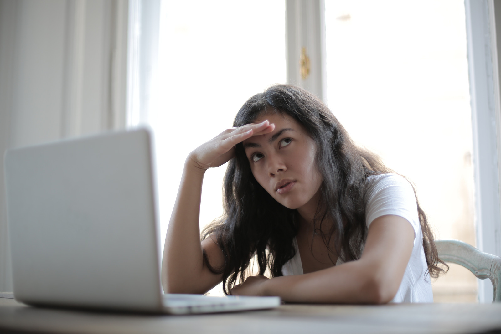 Eine junge Frau sitzt vor einem Laptop und schaut verzweifelt in die Ferne.